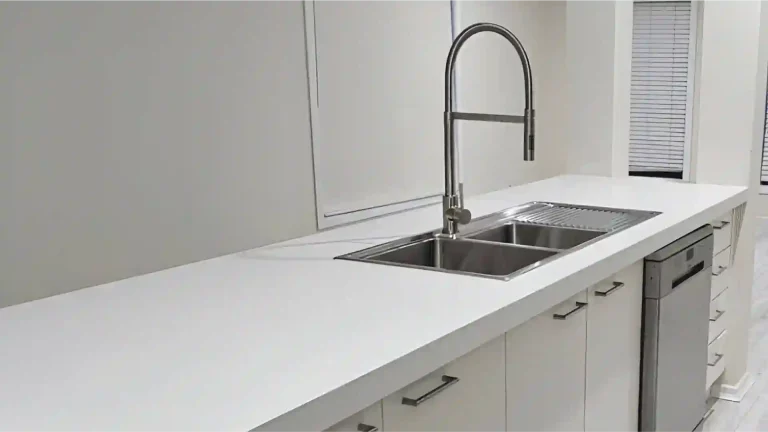Renovated kitchen featuring white countertop, stainless steel sink with high-arc faucet, built-in dishwasher, and white cabinetry. A sleek, minimalist design with natural light.