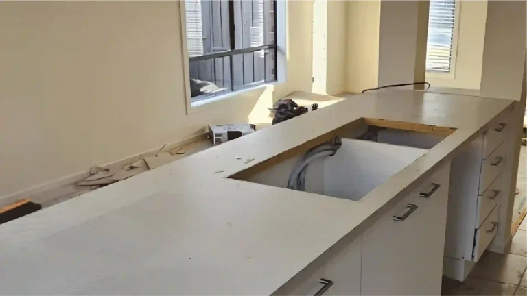 Kitchen under renovation with white countertop, open cabinetry, tools, and construction materials, pre-installation phase with natural light and protective flooring.